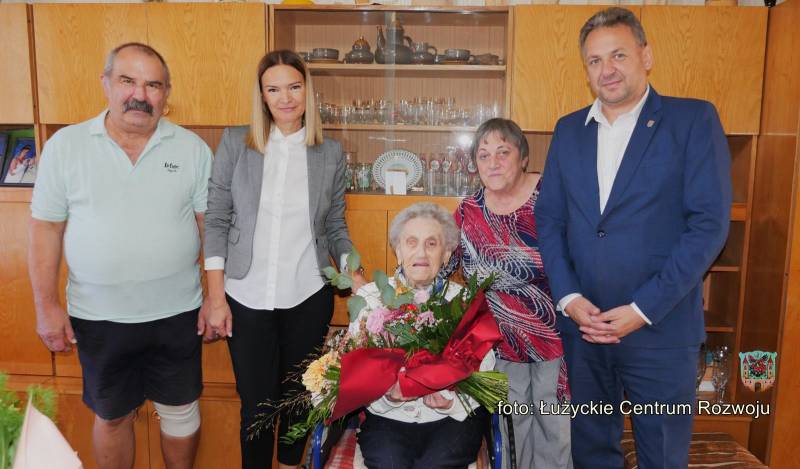 Pięć osób pozuje do zdjęcia, w środku starsza kobieta na wózku inwalidzkim trzymająca bukiet kwiatów, obok niej stoją dwie kobiety i dwóch mężczyzn, w tle klasyczna meblościanka